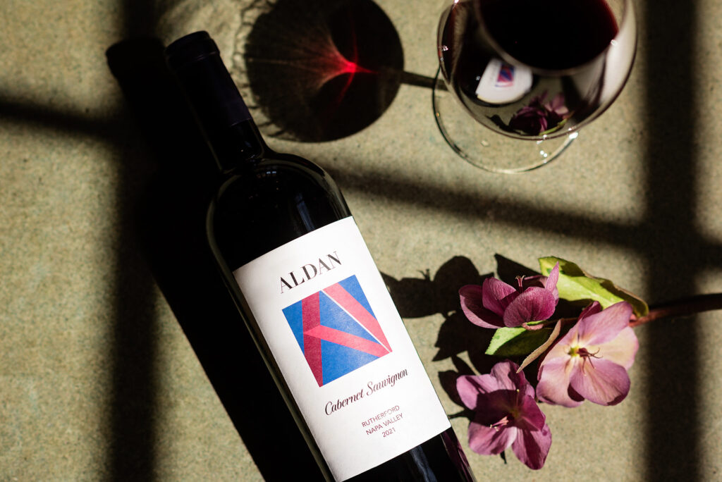 overheard view of ALDAN Wines Cabernet bottle laying flat beside a purple flower and filled glass